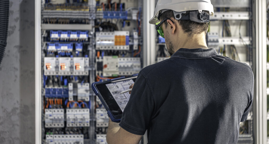 Man, an electrical working in a switchboard with fuses, uses a tablet.